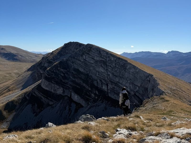 Un escursionista su un prato d'alta quota davanti a una parete di roccia stratificata, sotto un cielo sereno
