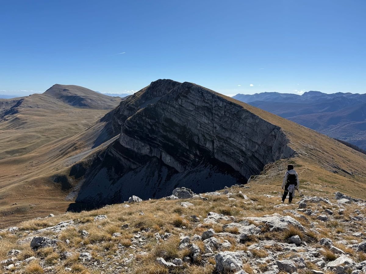 Il Balzo delle Fate: un enorme strapiombo di roccia a strati sulla cresta, con una persona che cammina sul bordo e le montagne del Parco sullo sfondo
