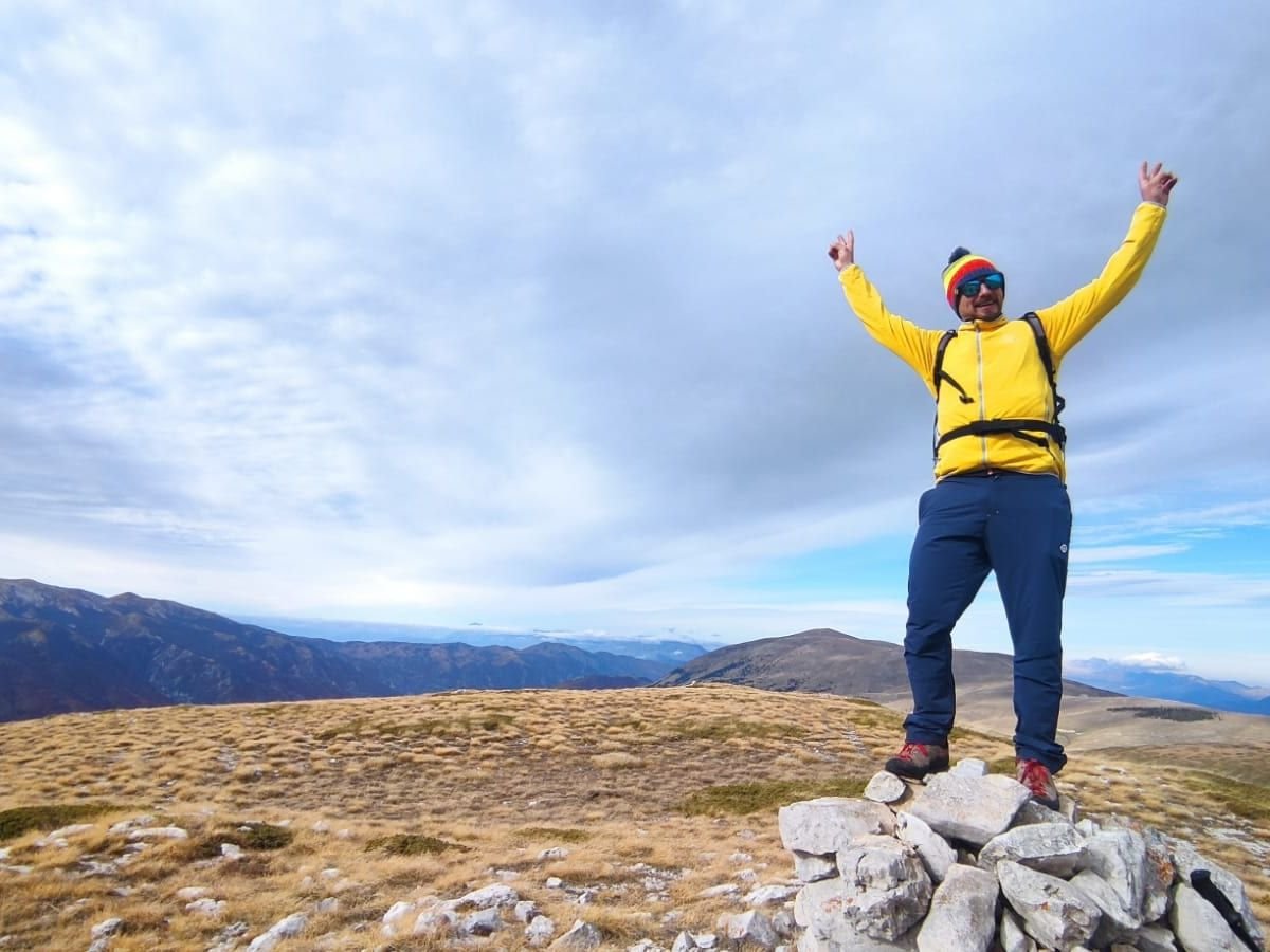 Un escursionista con la giacca gialla alza le braccia in cima alla Serra Sparvera, su un cumulo di pietre, con una cresta di erba secca dietro