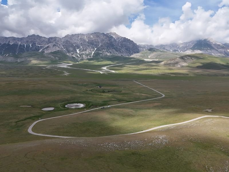 Campo Imperatore dal drone: l'altopiano verde attraversato da una strada a curve, con le montagne del Gran Sasso sotto le nuvole