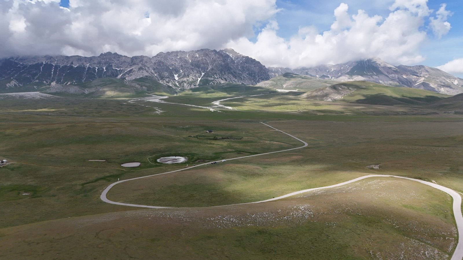 Campo Imperatore dal drone: l'altopiano verde attraversato da una strada a curve, con le montagne del Gran Sasso sotto le nuvole