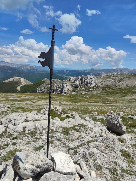 Un'asta di ferro con una bandierina sagomata su un mucchio di pietre, e dietro i pascoli e le rocce bianche della cresta delle Ciminiere
