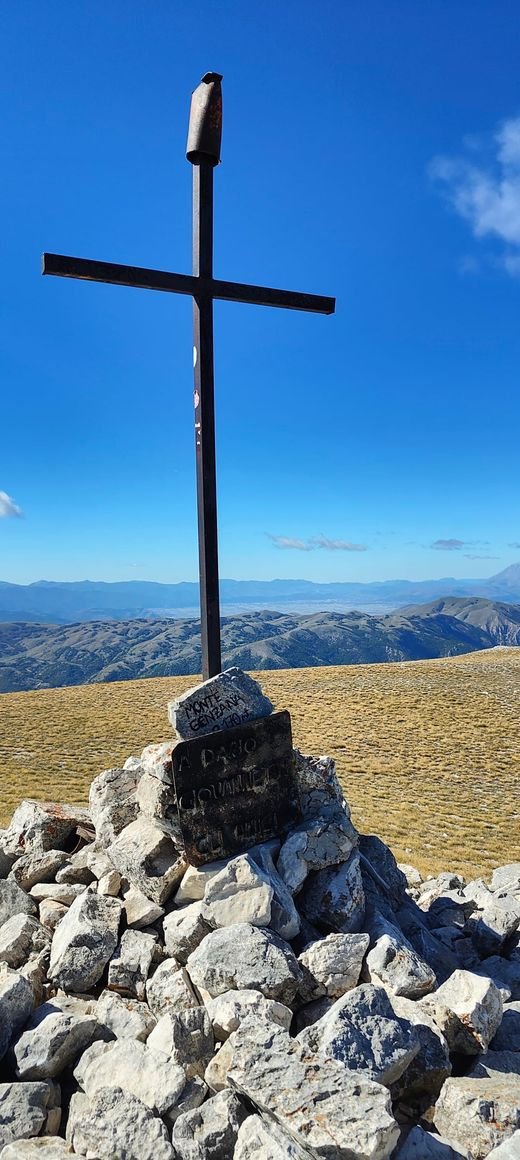 La croce di ferro sulla cima del Monte Genzana, su un cumulo di pietre con una pietra incisa e una targa, sopra i pascoli gialli e le montagne lontane