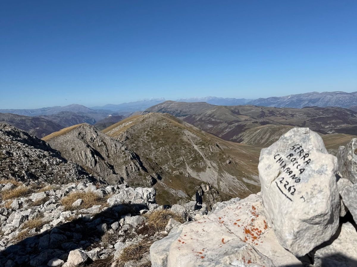 Il sasso di cima con la scritta a mano 'Serra Rocca Chiarano 2262', su un cumulo di pietre chiare, e dietro le creste e le valli del Parco sotto un cielo blu