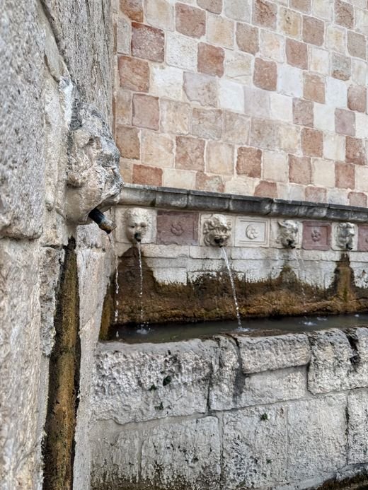 Una vasca di pietra della Fontana delle 99 Cannelle, con le cannelle che versano acqua e il muro a scacchi bianchi e rosa