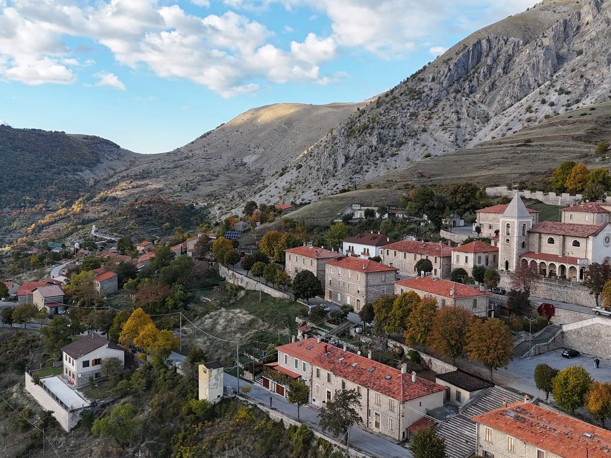 Frattura vista dal drone: case con i tetti rossi e un campanile tra gli alberi d'autunno, con i pendii spogli della montagna dietro