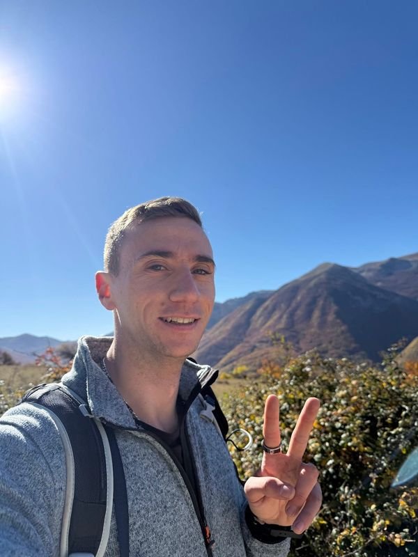 Giulio smiling and making a peace sign, backpack on, in front of autumn mountains