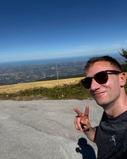 Giulio in un selfie, con gli occhiali da sole e due dita alzate, su un altopiano con il mare all'orizzonte