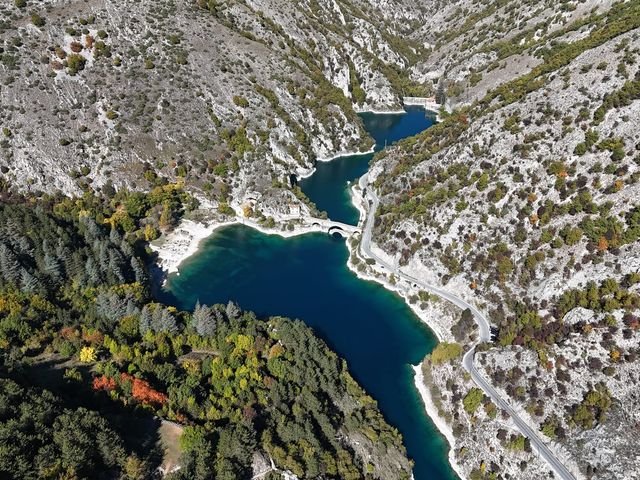 Il Lago di San Domenico visto dal drone: acqua blu scuro nella gola tra pareti di roccia, con il ponte ad arco, l'eremo sulla riva e la strada che costeggia il lago