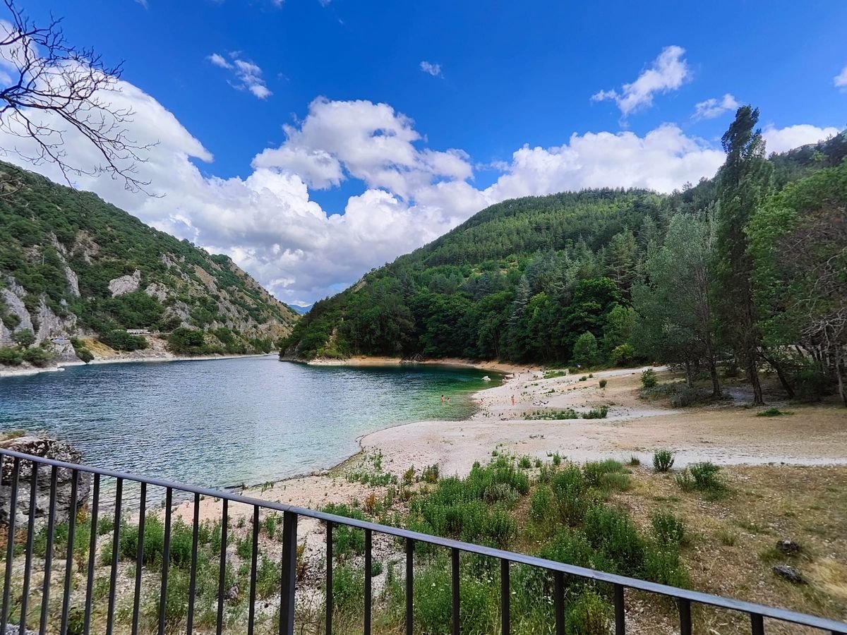 La spiaggetta di ciottoli del Lago di San Domenico, con l'acqua verde e i boschi sulle due sponde, vista da una balaustra di ferro