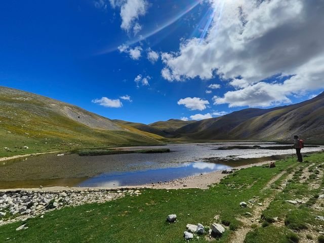Il Lago di Pantaniello con poca acqua, tra pendii di erba, con un escursionista fermo sulla riva e un cielo blu con grandi nuvole
