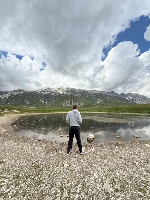 Una persona in piedi accanto al piccolo Lago Pietranzoni, con le nuvole riflesse nell'acqua e l'altopiano intorno