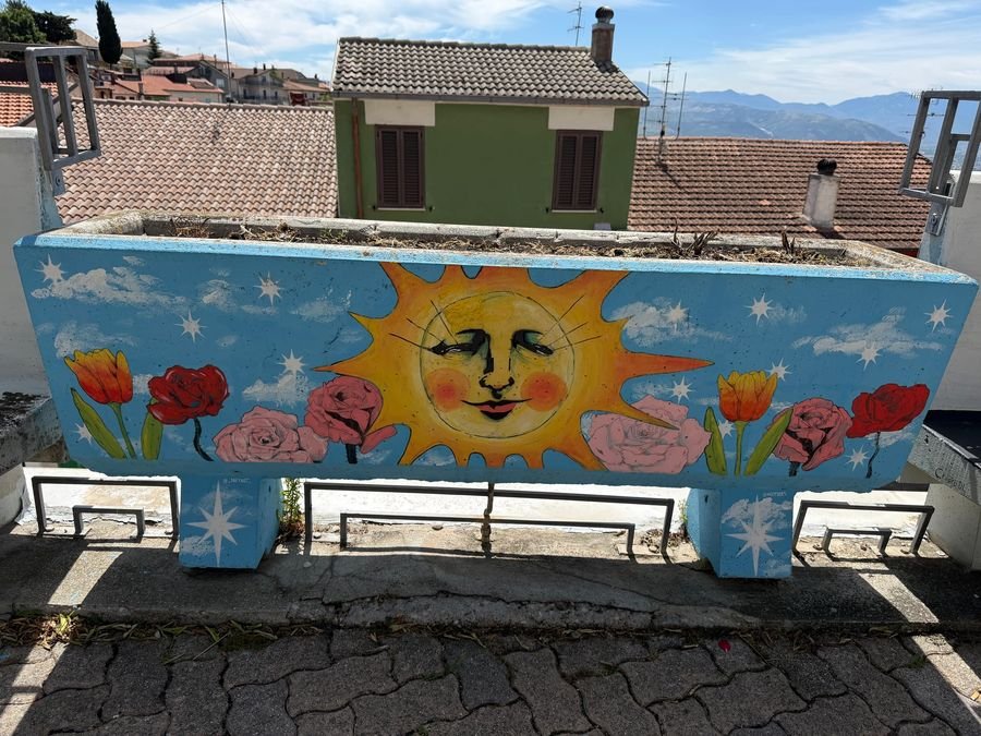 Una fioriera dipinta con un grande sole sorridente e dei fiori rossi, con i tetti di Aielli e le montagne dietro