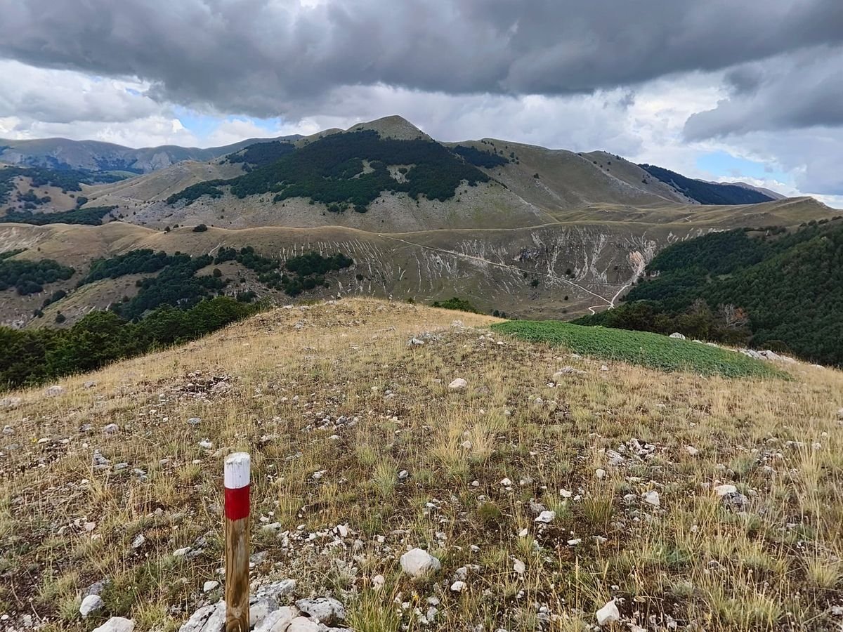 Un paletto bianco e rosso del CAI su un crinale di erba e sassi, davanti ai Monti Marsicani sotto le nuvole