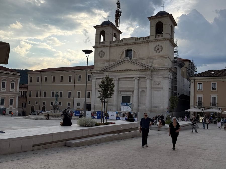 Piazza Duomo all'Aquila: la piazza ampia con la facciata del Duomo e alcuni passanti, sotto un cielo nuvoloso