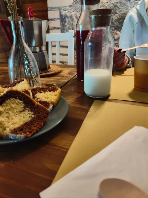 Una colazione sul tavolo di legno: una fetta di ciambellone, una caraffa di latte, una moka e una bottiglia di succo