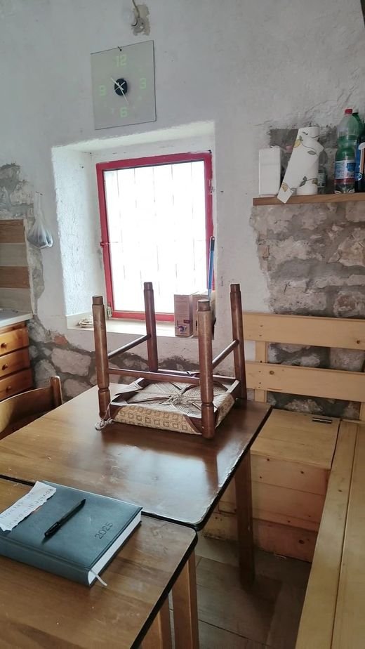 Un tavolo di legno con un quaderno sopra, le sedie capovolte e una finestra dagli infissi rossi nel muro di pietra