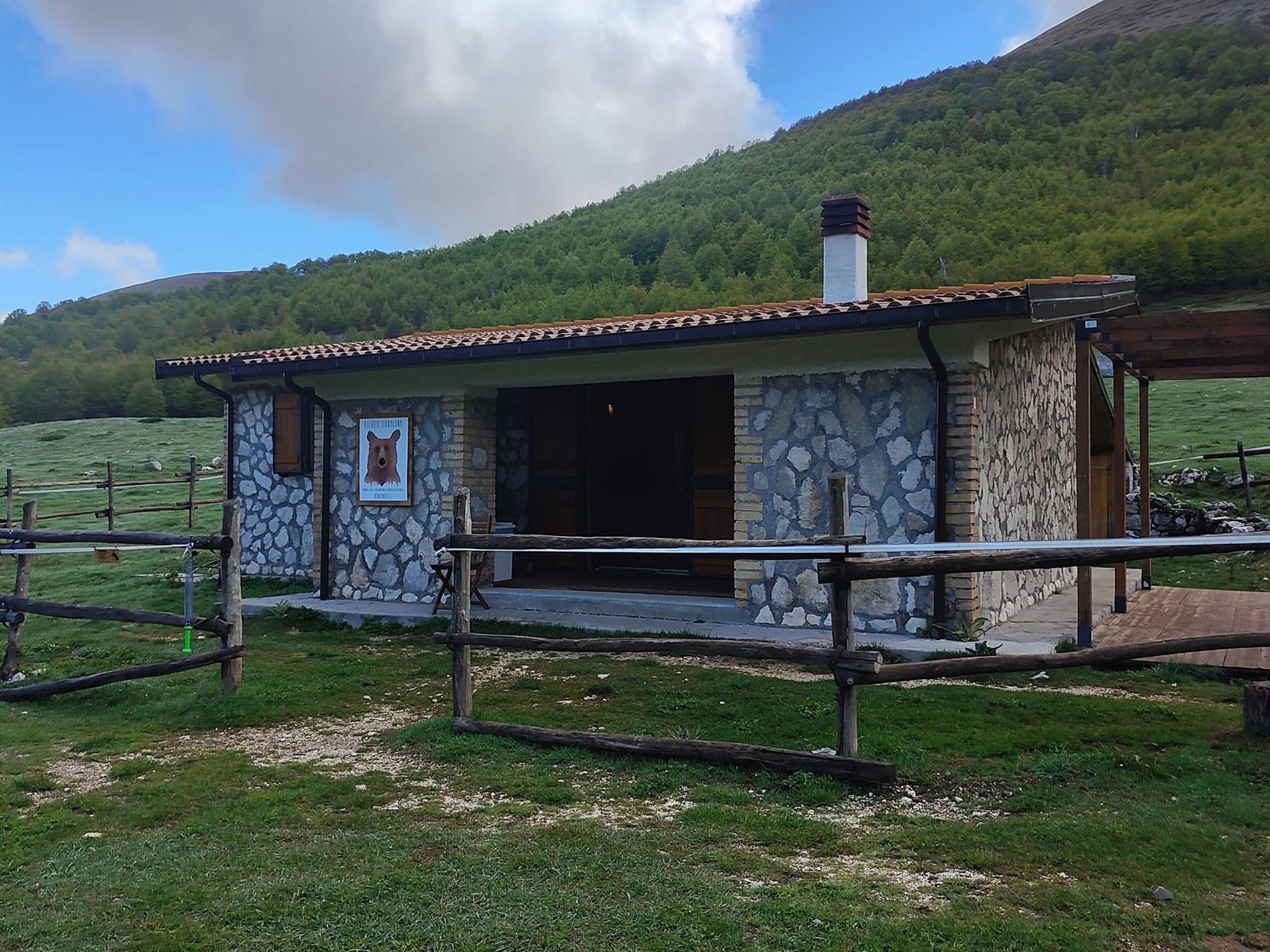 Il Rifugio di Terraegna: casetta di pietra con il camino e uno steccato di legno, su un prato di montagna sotto grandi nuvole bianche