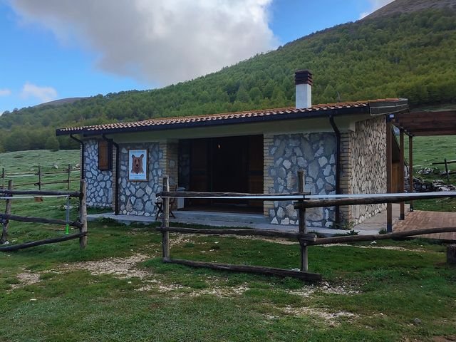 Il Rifugio di Terraegna: casetta di pietra con il camino e uno steccato di legno, su un prato di montagna sotto grandi nuvole bianche