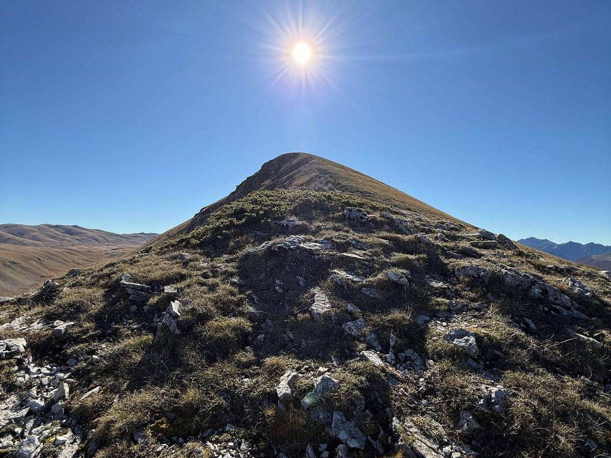 La cresta di Serra Rocca Chiarano con rocce e ginepri in primo piano e la cima a punta contro il cielo, con il sole proprio sopra