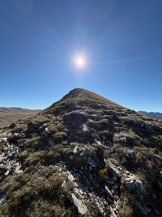 La cresta di Serra Rocca Chiarano con rocce e ginepri in primo piano e la cima a punta contro il cielo, con il sole proprio sopra