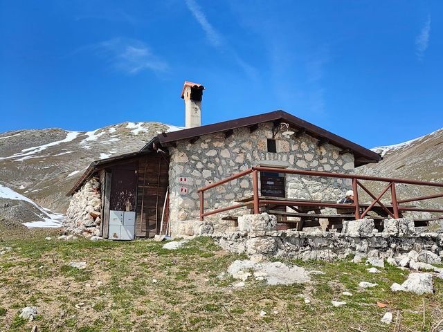 Lo Stazzo del Campo: una casetta di pietra con il camino e una staccionata di legno, su un prato in quota con la neve sulle montagne dietro