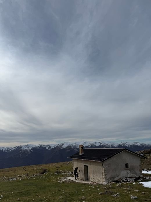 Lo Stazzo del Genzana, una casetta di pietra con il tetto scuro e il camino, su un prato con la neve a chiazze e montagne innevate dietro
