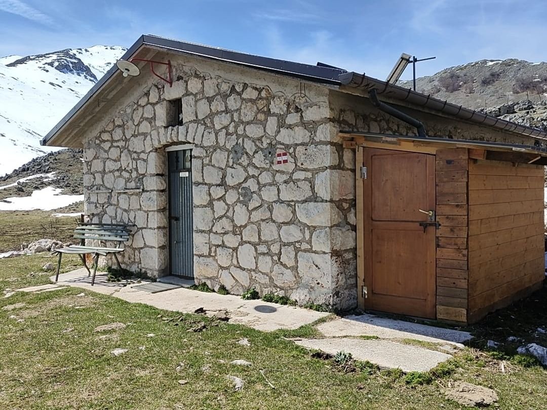 Lo Stazzo di Campo Rotondo: una casetta di pietra con una parte in legno, su un prato in quota con la neve sulle montagne dietro