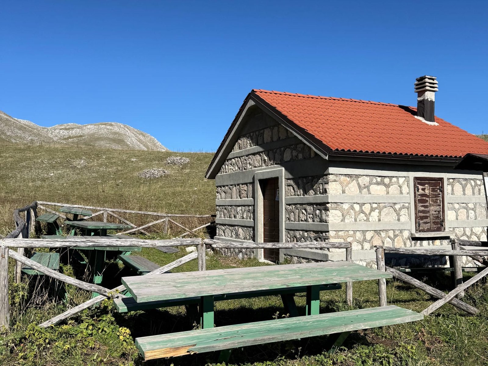 Lo Stazzo La Montagnola: casetta di pietra con il tetto rosso e il camino, con due tavoli da picnic di legno sul prato davanti