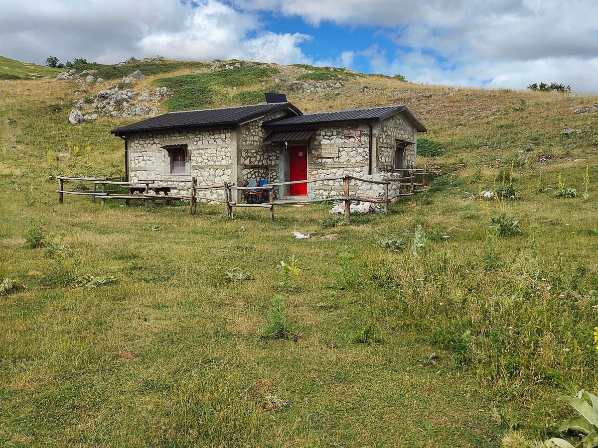 Il Rifugio Pagliaccio di Preccia: casetta di pietra con il tetto scuro e la porta rossa, circondata da una staccionata di legno, su un prato