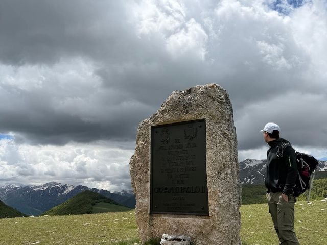 La Stele del Papa, un grande masso con una targa di bronzo, su un prato di montagna, con un escursionista che la guarda e cime lontane sotto le nuvole