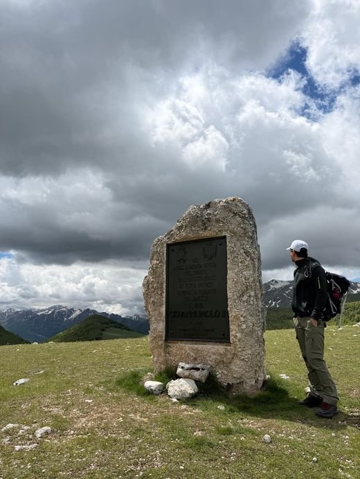 La Stele del Papa, un grande masso con una targa di bronzo, su un prato di montagna, con un escursionista accanto e cime lontane sotto le nuvole
