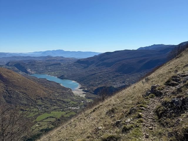 Il sentiero di sassi e terra su un pendio d'erba, con il Lago di Barrea e la valle in fondo sotto un cielo azzurro