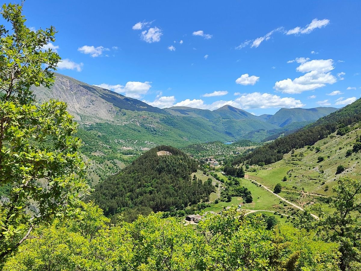 Una valle verde vista dall'alto, con un lago in fondo e le montagne dietro, sotto un cielo blu
