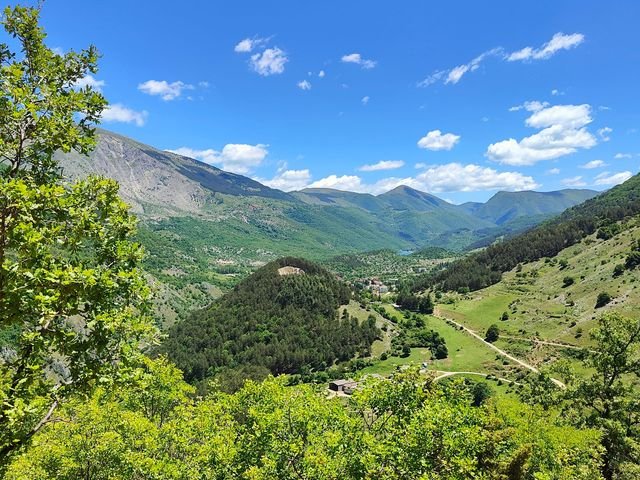 Una valle verde vista dall'alto, con un lago in fondo e le montagne dietro, sotto un cielo blu