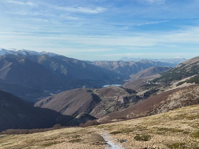 Il Lago di Scanno in fondo alla valle, tra montagne con la neve sulle creste, e un sentiero in mezzo all'erba in primo piano