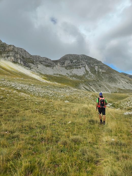 Un escursionista con lo zaino cammina sull'erba verso un'alta parete di roccia grigia, la Serra di Rocca Chiarano