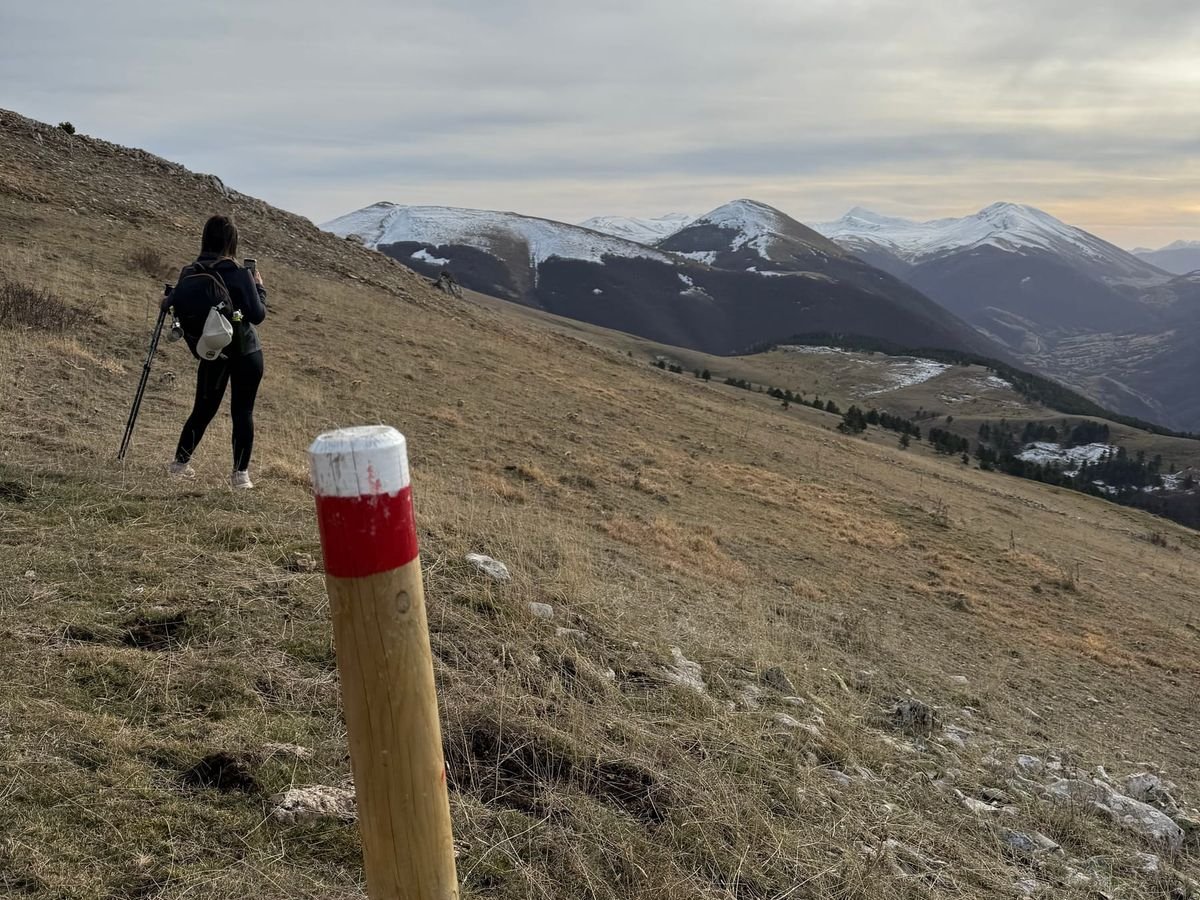 Un palo di legno con il segno bianco e rosso del sentiero su un pendio d'erba, un'escursionista più avanti e montagne con un po' di neve dietro